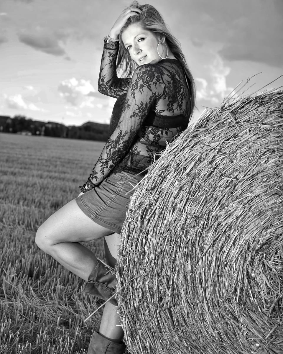 A woman in a lace top leaning against a round straw bale on the stubble