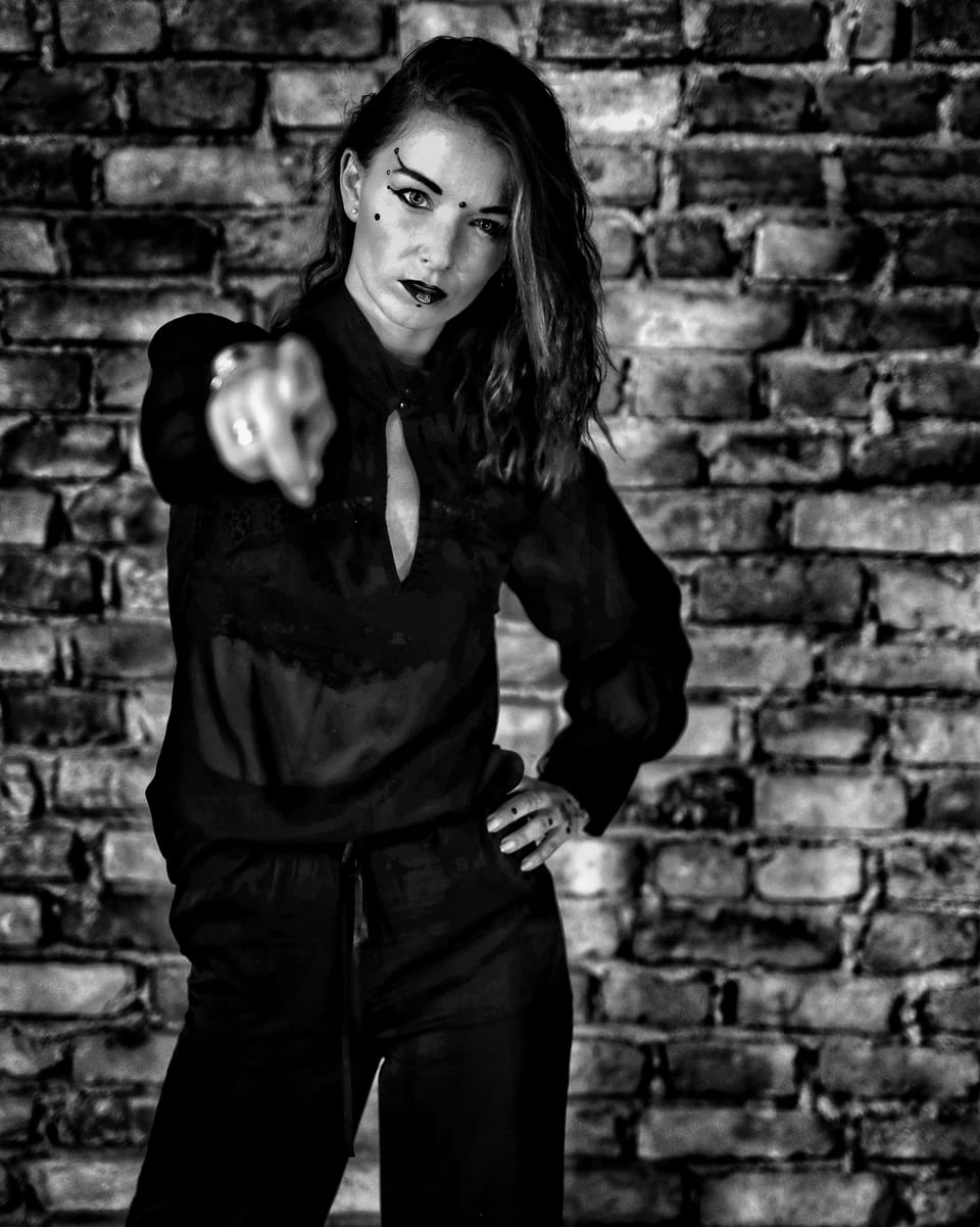 A woman in black at a brick wall pointing a finger straight into the lens