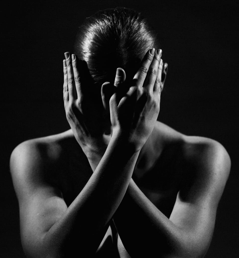 Crossed arms and palms across the face, only the shoulders emerging from the dark