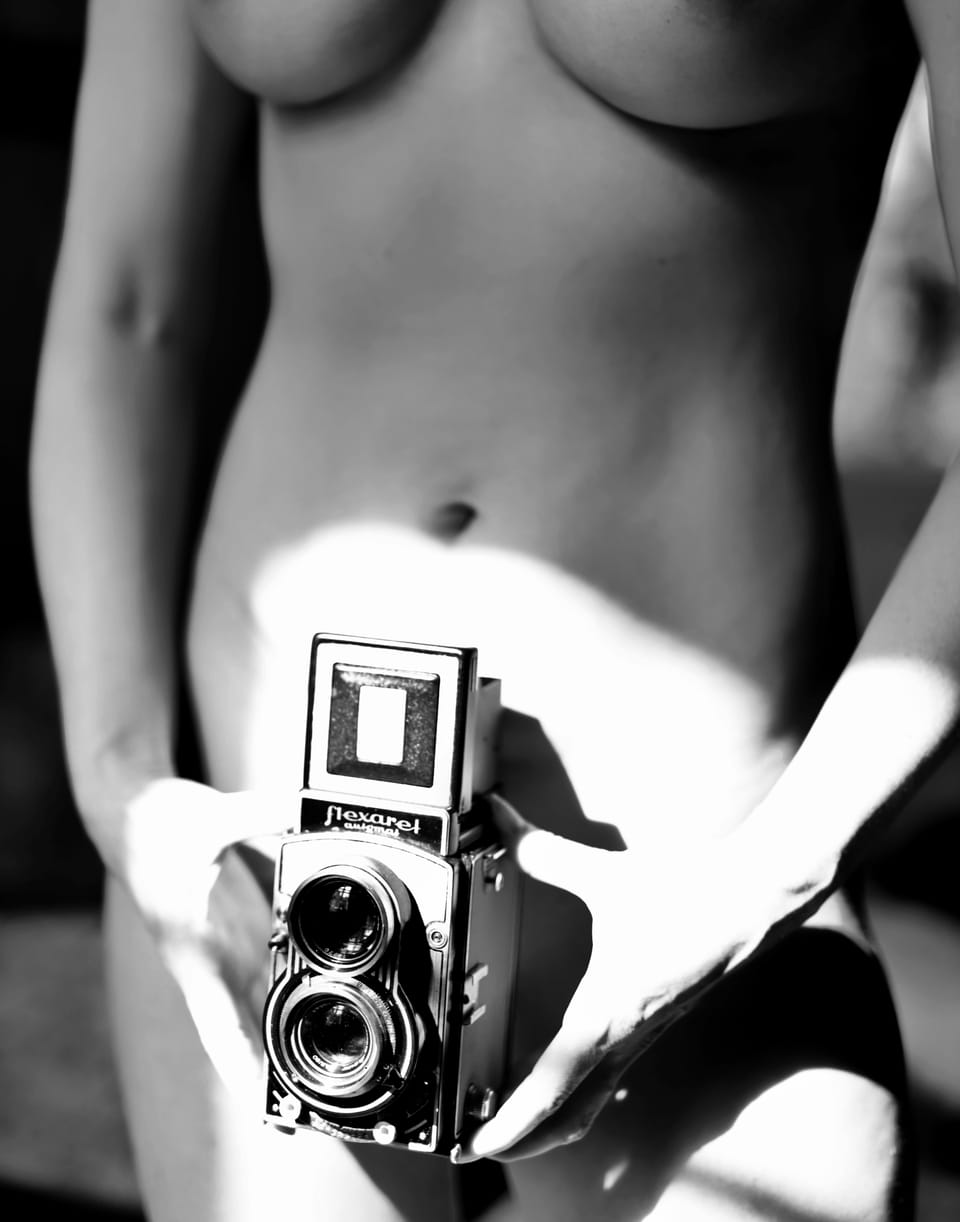 A black and white nude study, the torso holding a twin-lens Flexaret camera