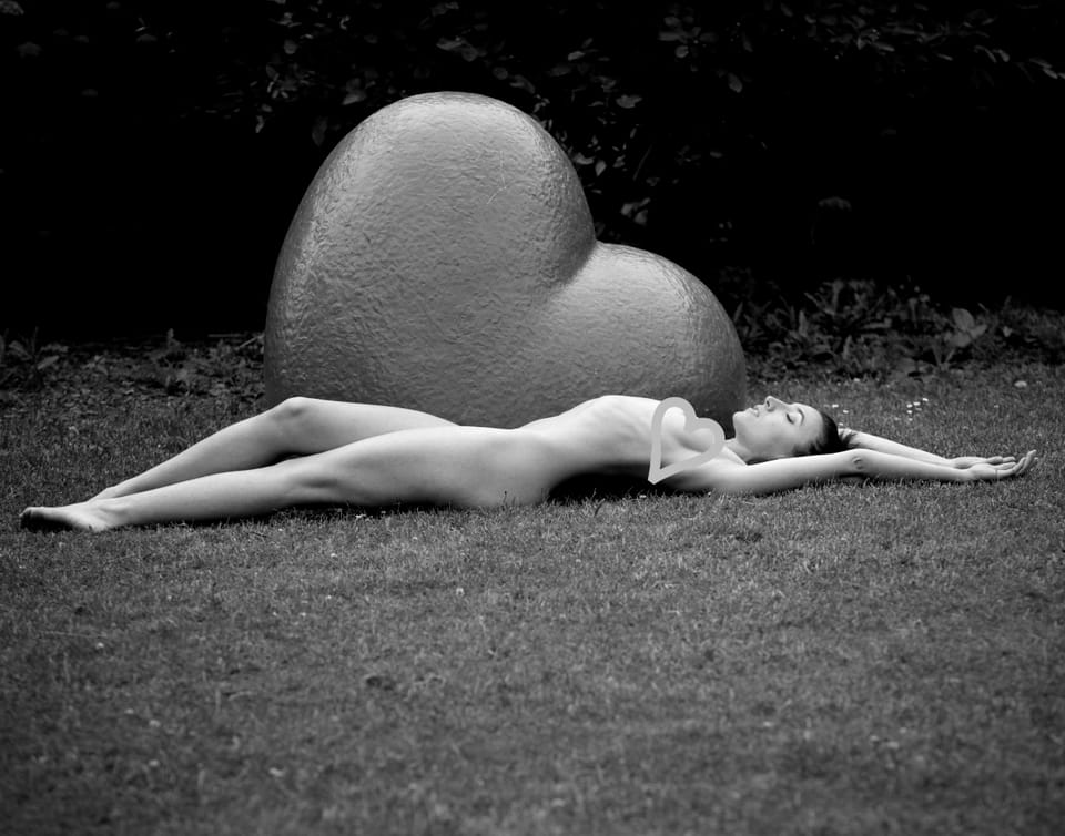 A nude lying on grass beside a stone sculpture shaped like a heart
