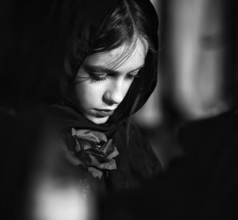 A young woman with her head lowered under a hood, deep darkness around