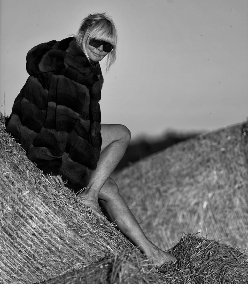 A woman in a fur coat and dark glasses seated on a straw bale above a field