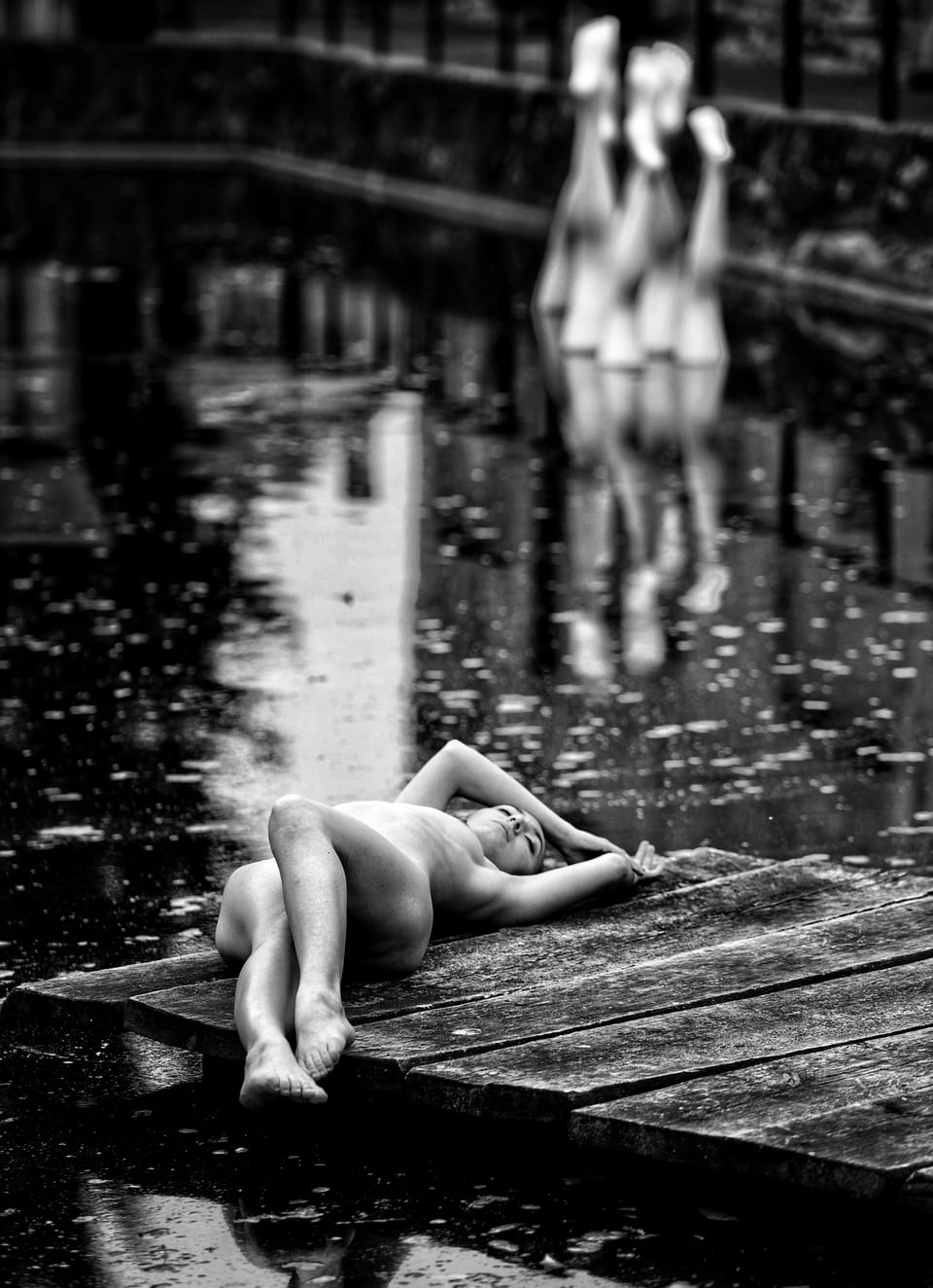 A nude reclining on a wooden pier in the rain, two blurred figures in the distance