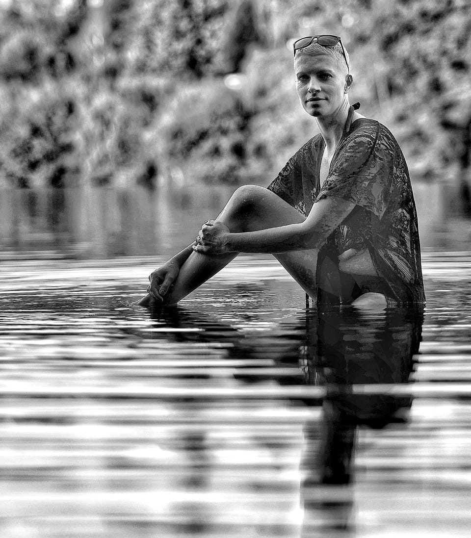 A figure seated in shallow water, the surface returning the whole image beneath