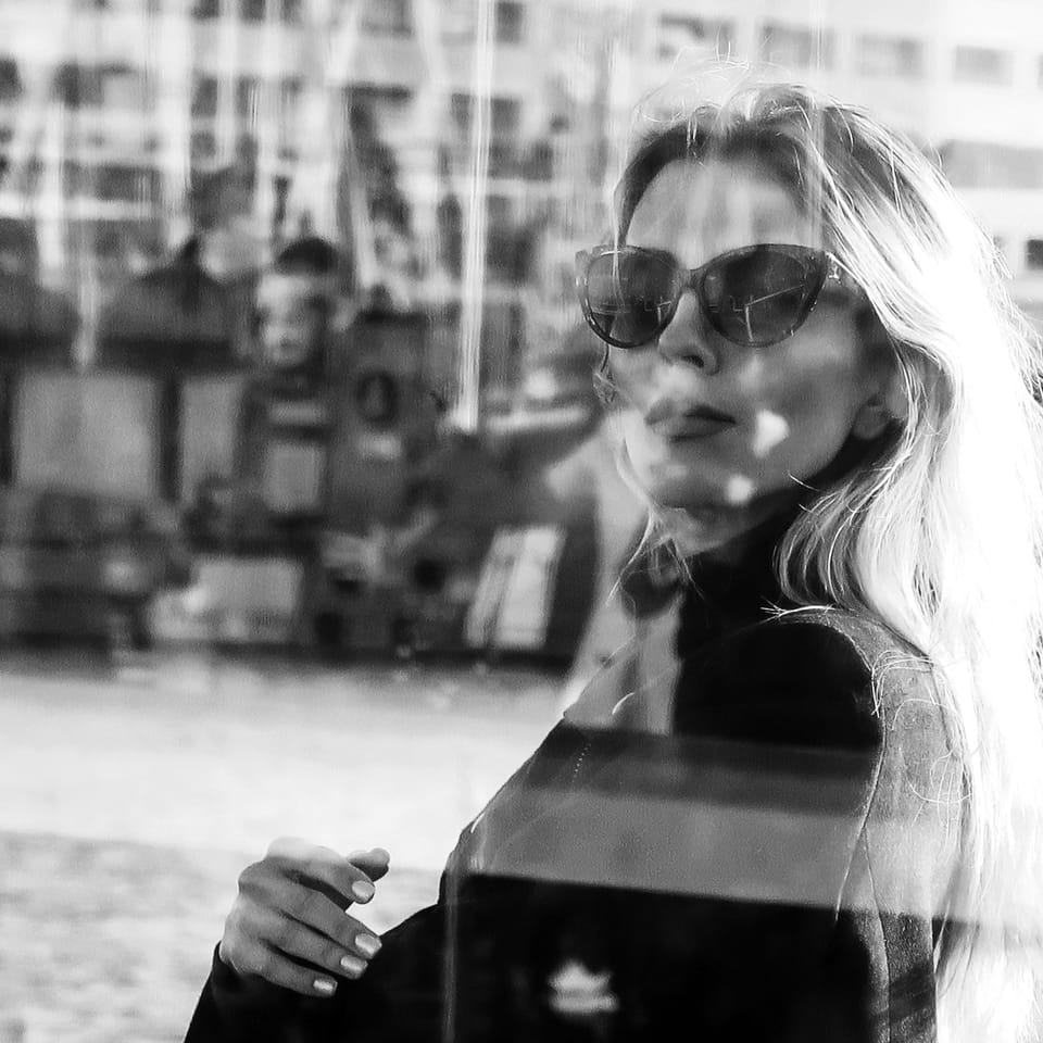 A woman in dark glasses behind glass, the town folding across her in the reflection