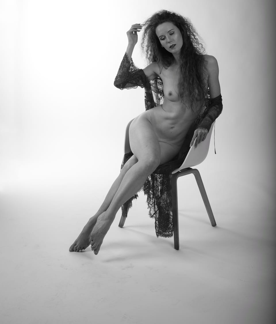 A woman on a chair against white, lace slipped down her arm