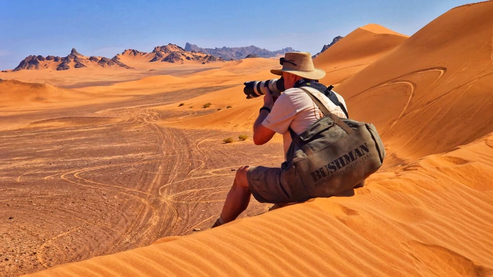 A photographer sits on the crest of an orange dune, lens aimed down into the valley, his bag propped in the sand