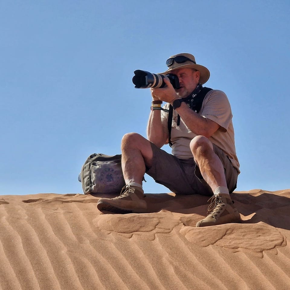 A photographer on the sand, a long telephoto to his eye, against a clean blue sky