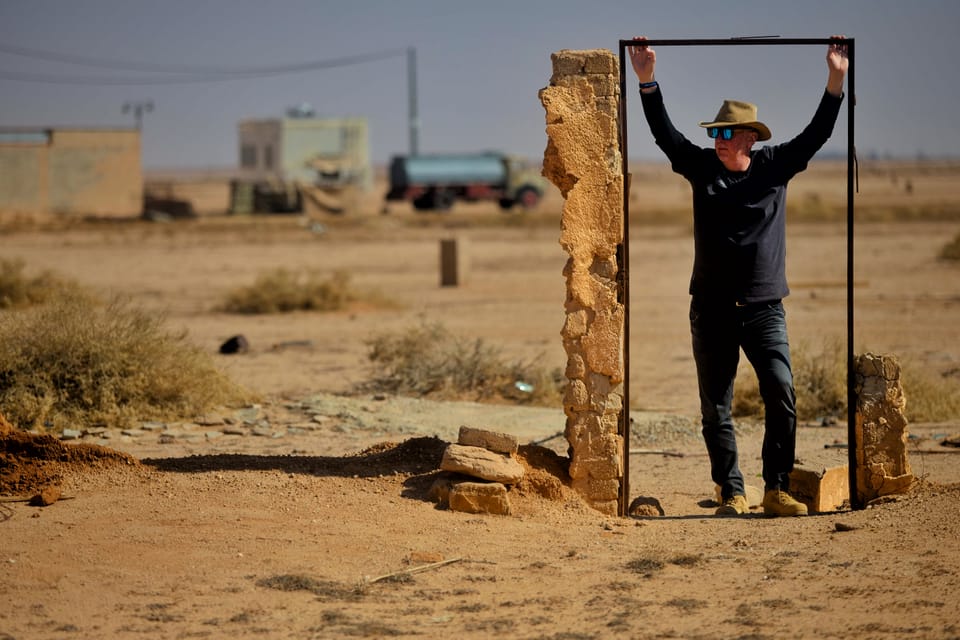 A man lifting a stone frame overhead, framing a piece of a desert village with it