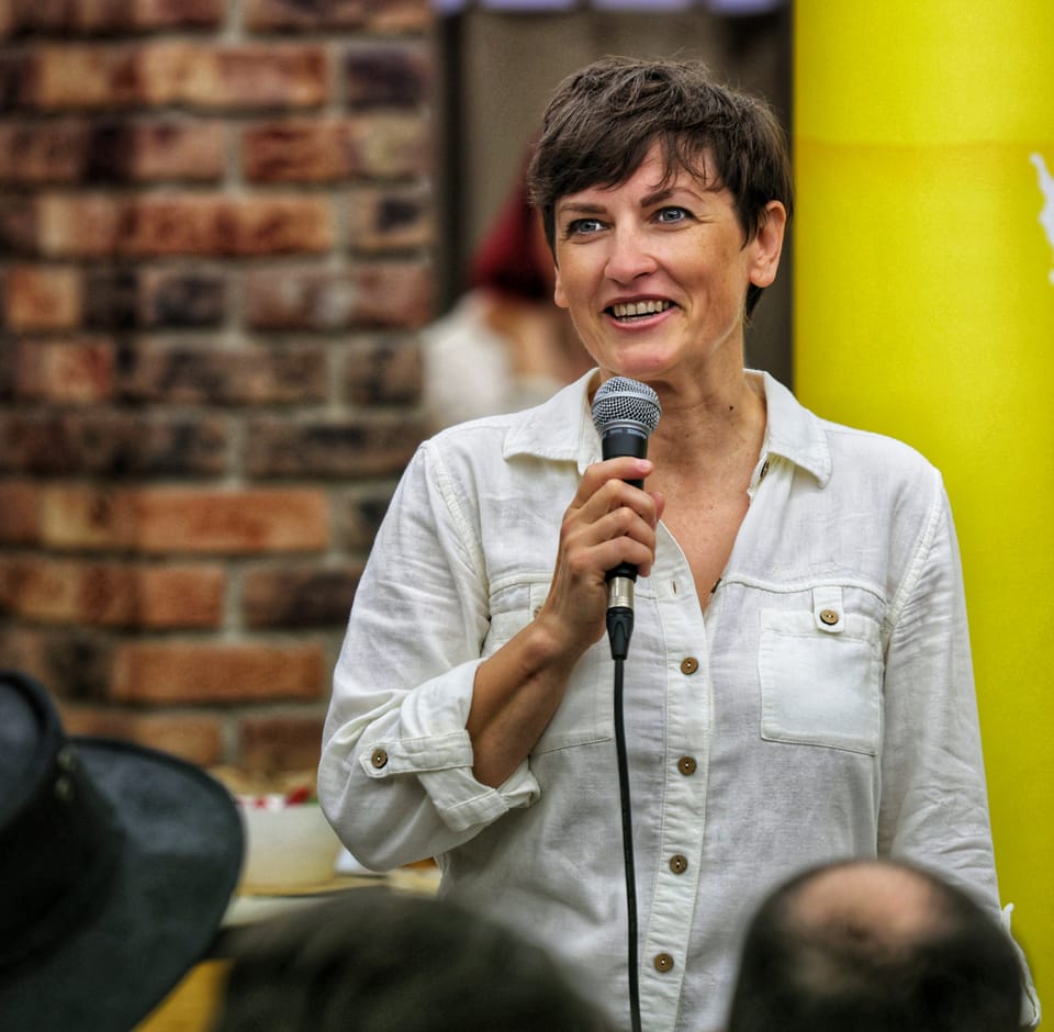 A woman speaking into a microphone, a brick wall and a blurred room behind