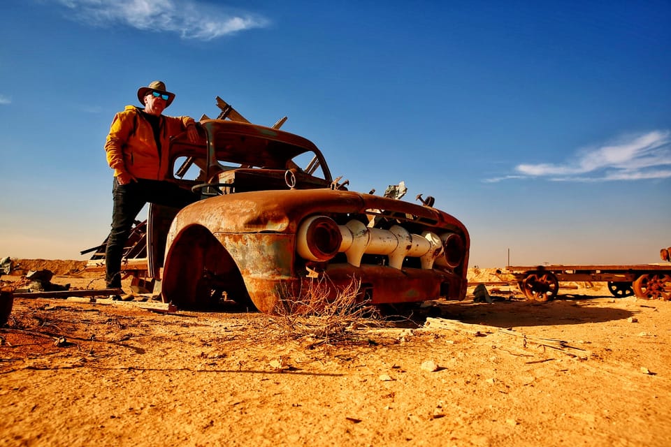 A figure standing on a rusted car wreck in the desert under a hard sun