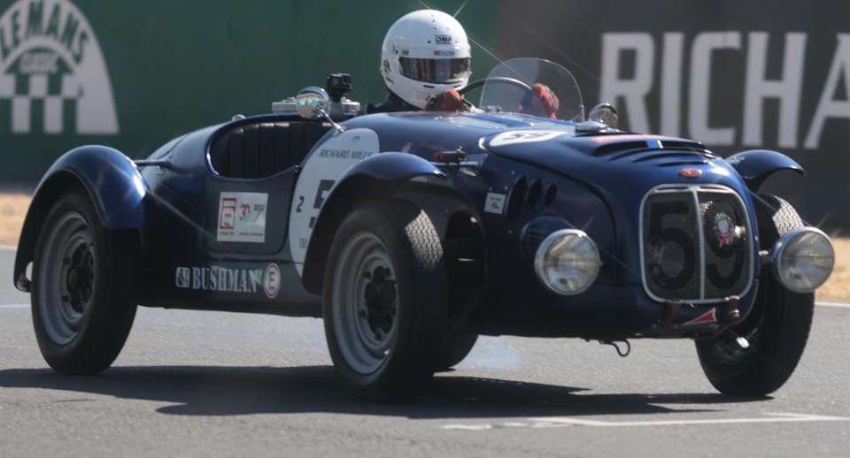 A dark blue historic roadster through a circuit bend, partner decals on its flank