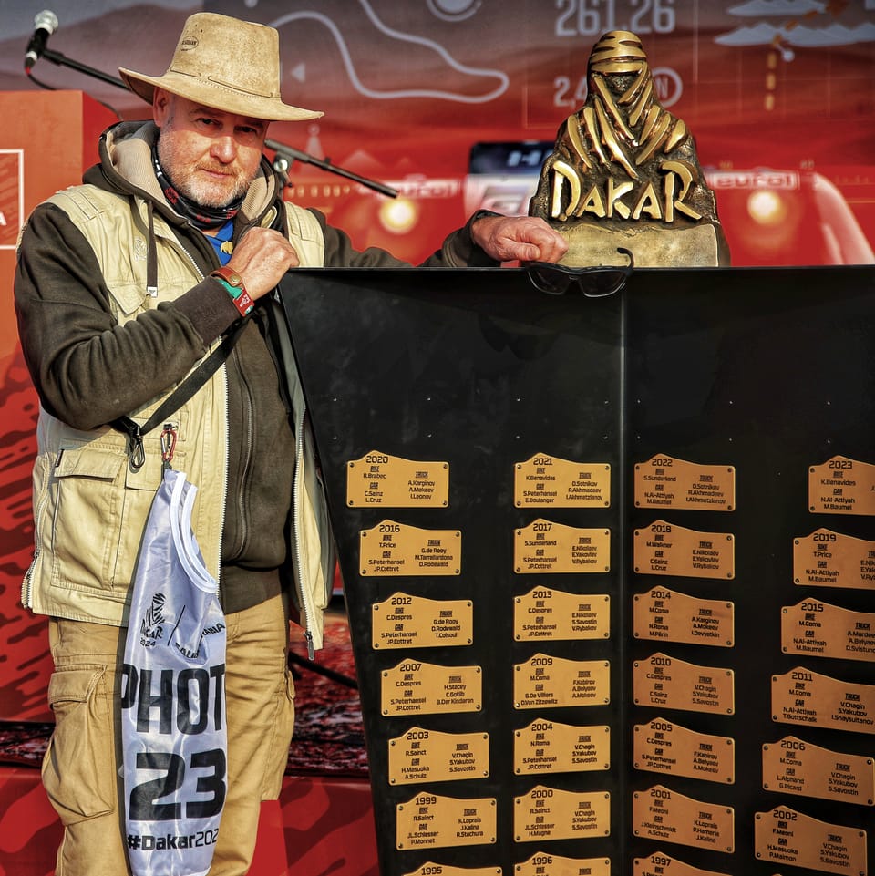 A photographer in a press bib beside a board of gold winners' plaques