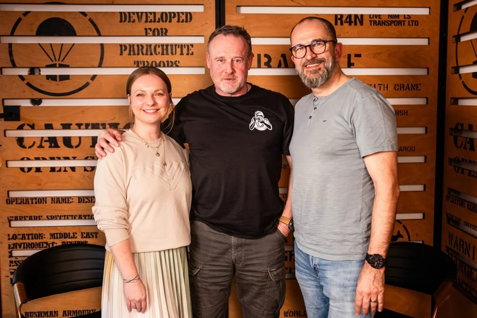Three people posing before a wall of crate boards stencilled with military lettering