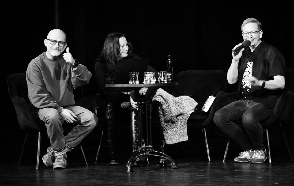 A black and white frame of an on-stage discussion, three people at a small table with a microphone