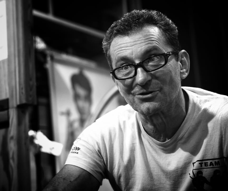 A man in glasses and a white shirt sitting against the light, photographs on the wall behind