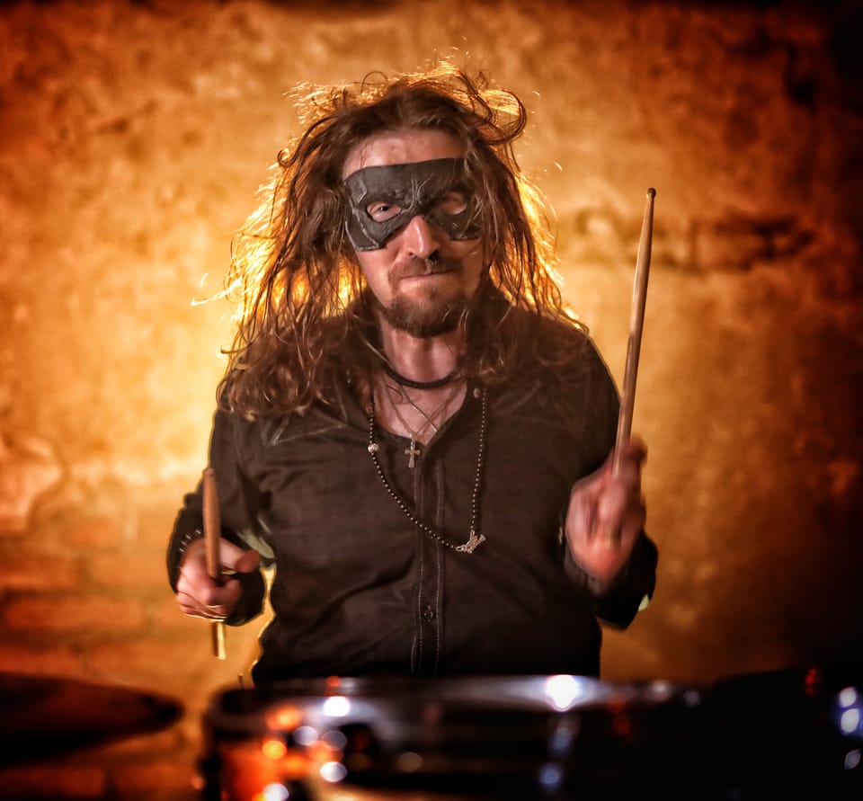 A drummer in a mask with sticks raised in warm spotlight
