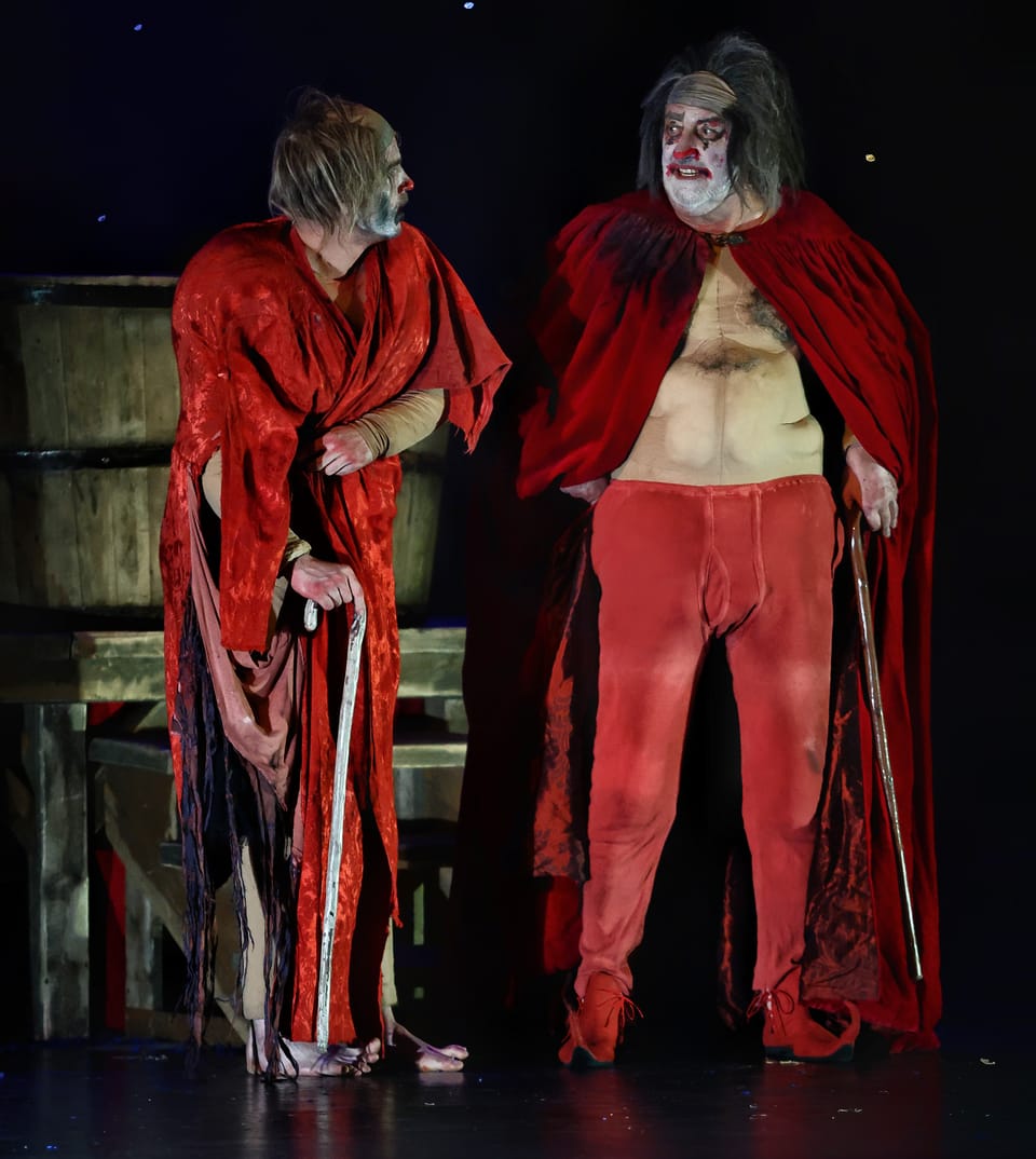 Two figures in ragged red cloaks with canes on a set of bare boards