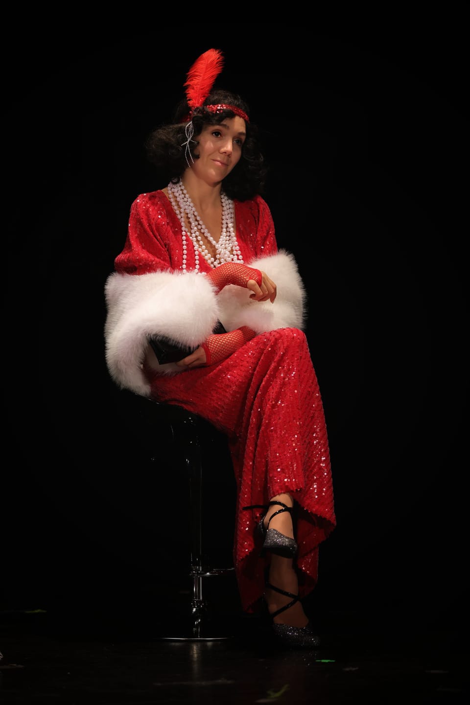 An actress in a red dress with a feather in her hair on a high stool