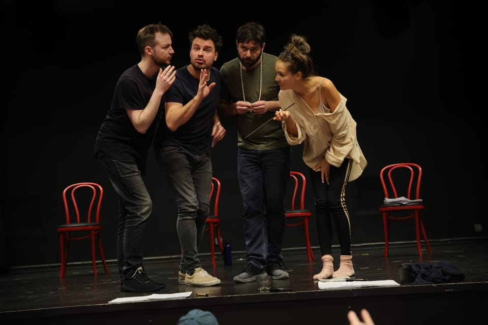 Four actors among red chairs, all turned toward a single point