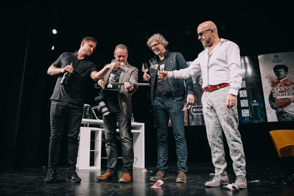 Four men on stage before a wall of posters, one handing something aside