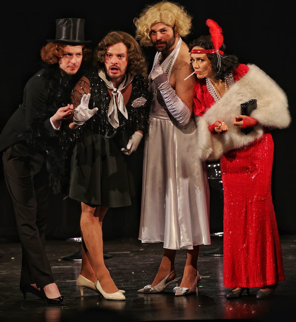 Five actors in period costume lined up, a woman in a red dress at the centre