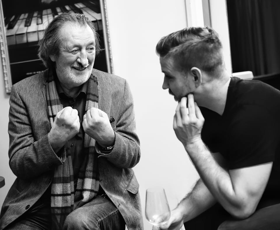 Two men facing each other over a glass, one laughing while the other watches
