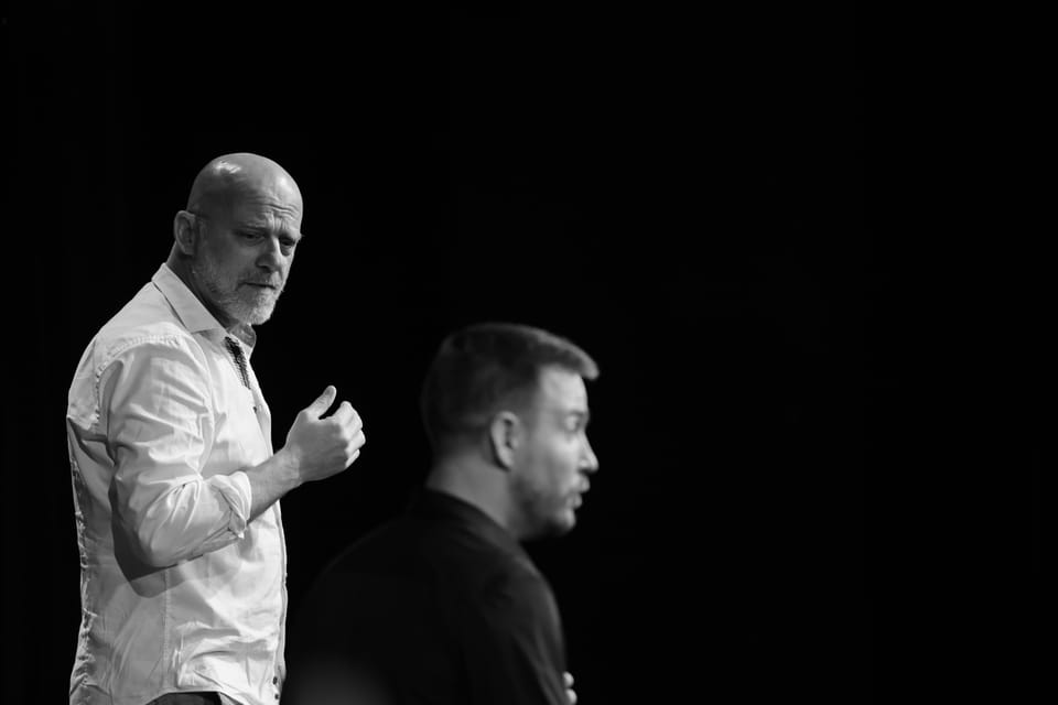 A man in a white shirt emerging from the dark, another with his back turned in front