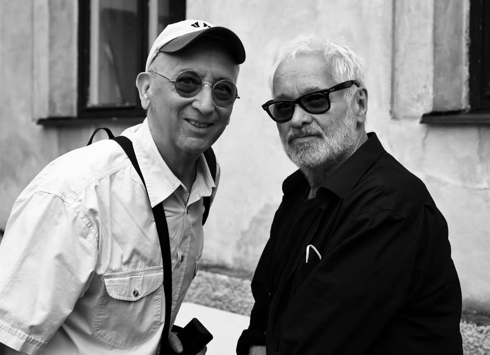 Two older men in glasses side by side at a wall, one in a cap