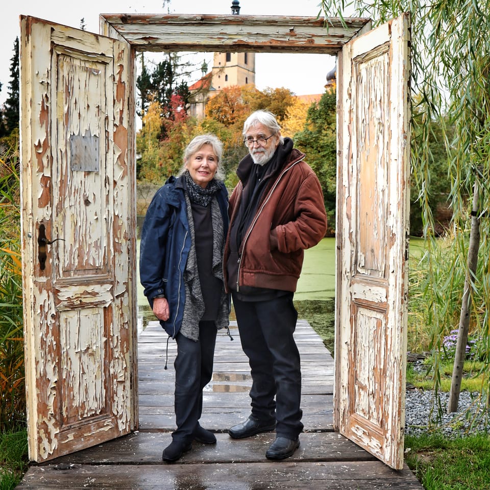 An older couple standing in a crumbling doorway, an autumn garden behind