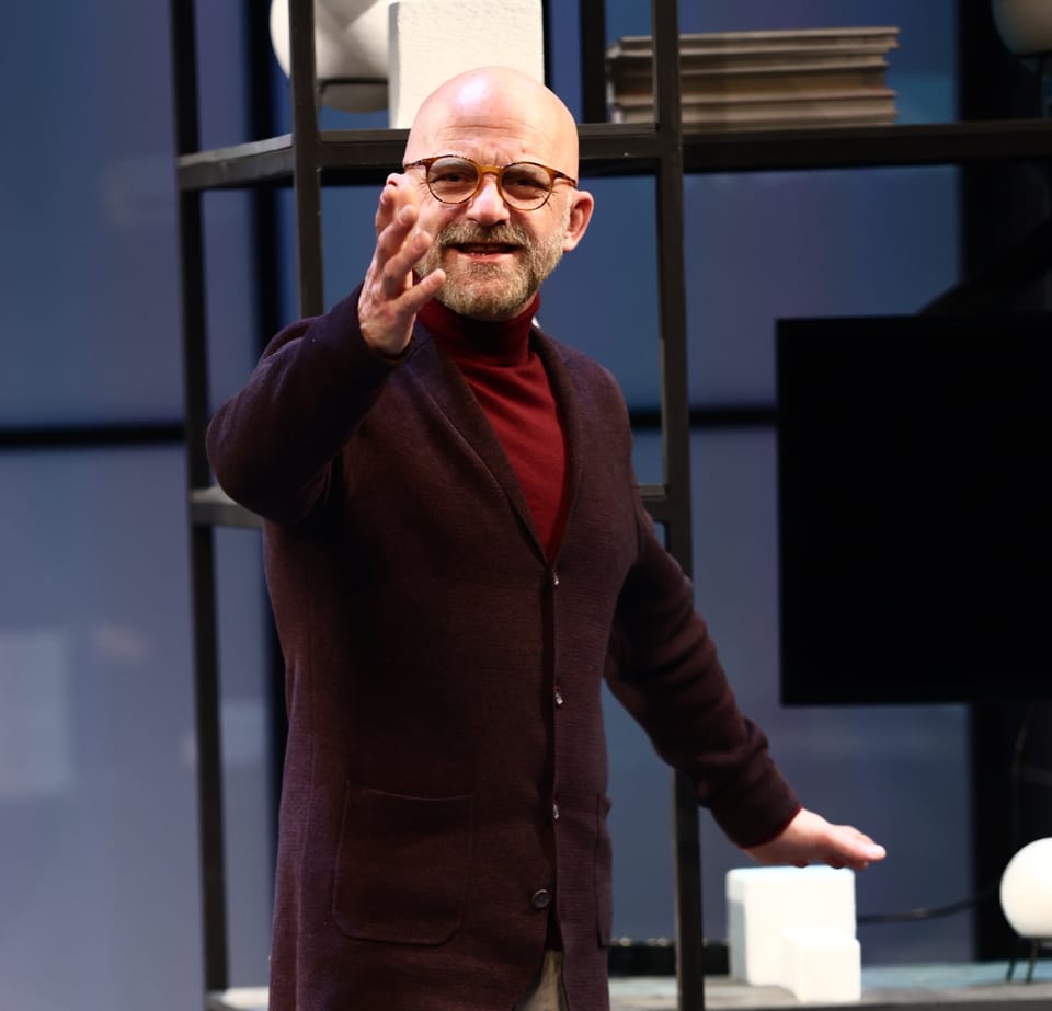 An actor with an open palm mid-line, a set of shelves behind him