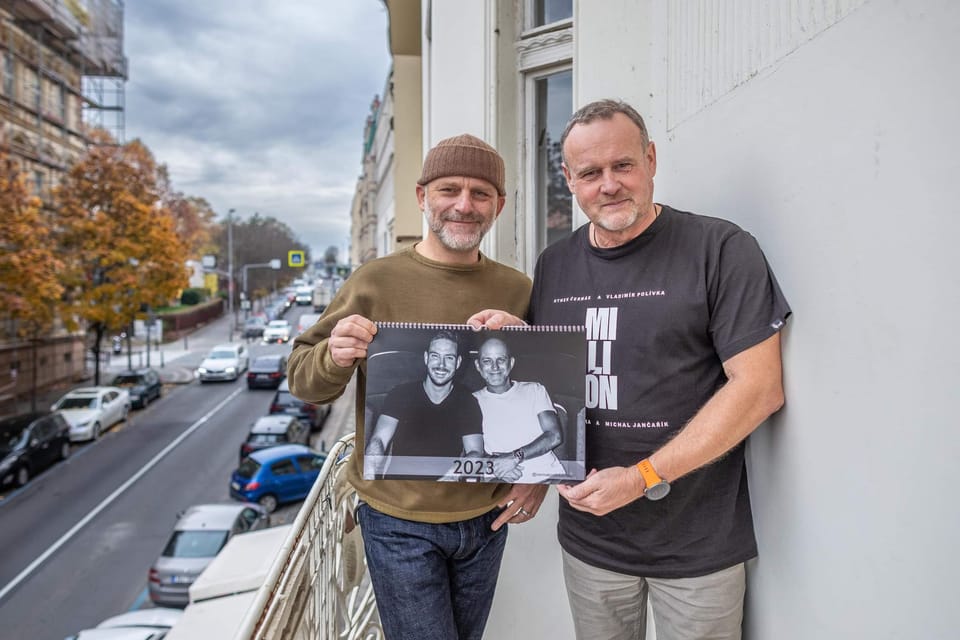 Two men holding a printed calendar between them, an autumn street behind