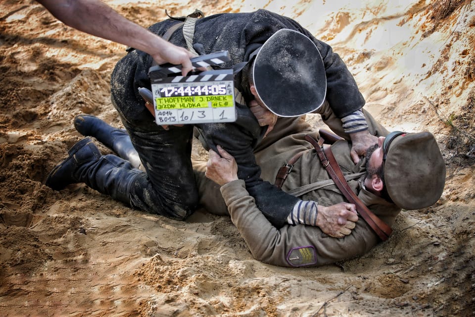 A clapperboard above two actors lying in the sand, both in period uniform