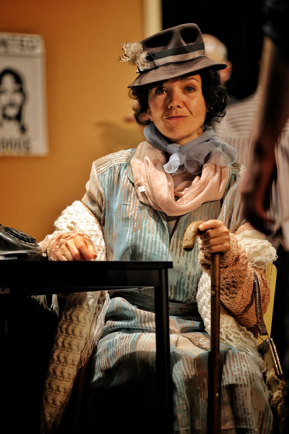 An actress in a hat and layered costume seated, a cane against her knee
