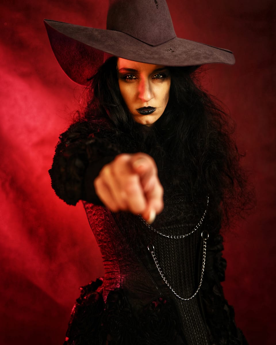 A figure in a wide hat and dark make-up pointing a finger into the lens