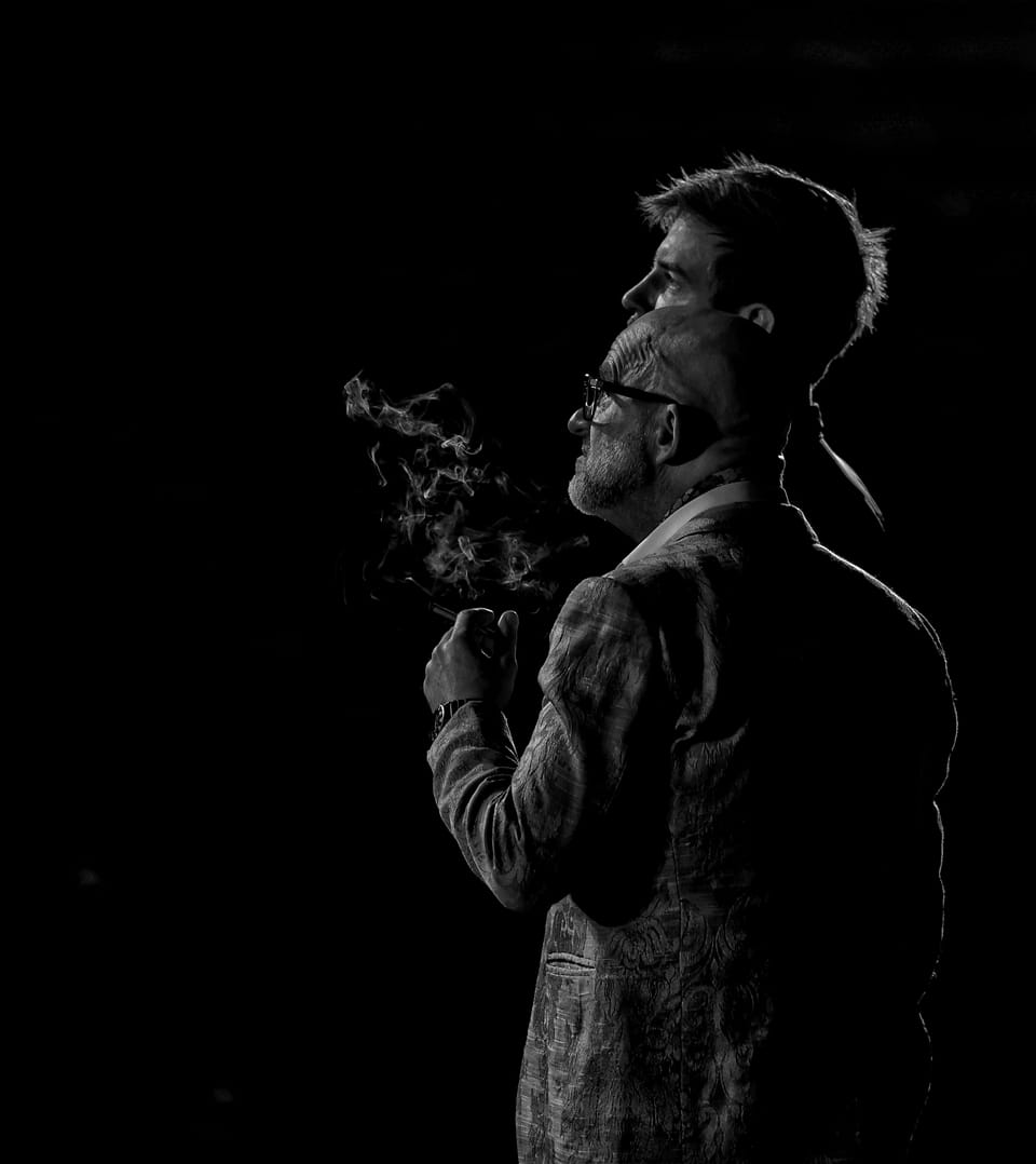 A man in profile breathing out smoke, another outlined in the dark behind him