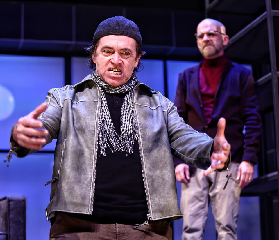 An actor in a leather jacket and cap with hands spread, another standing behind