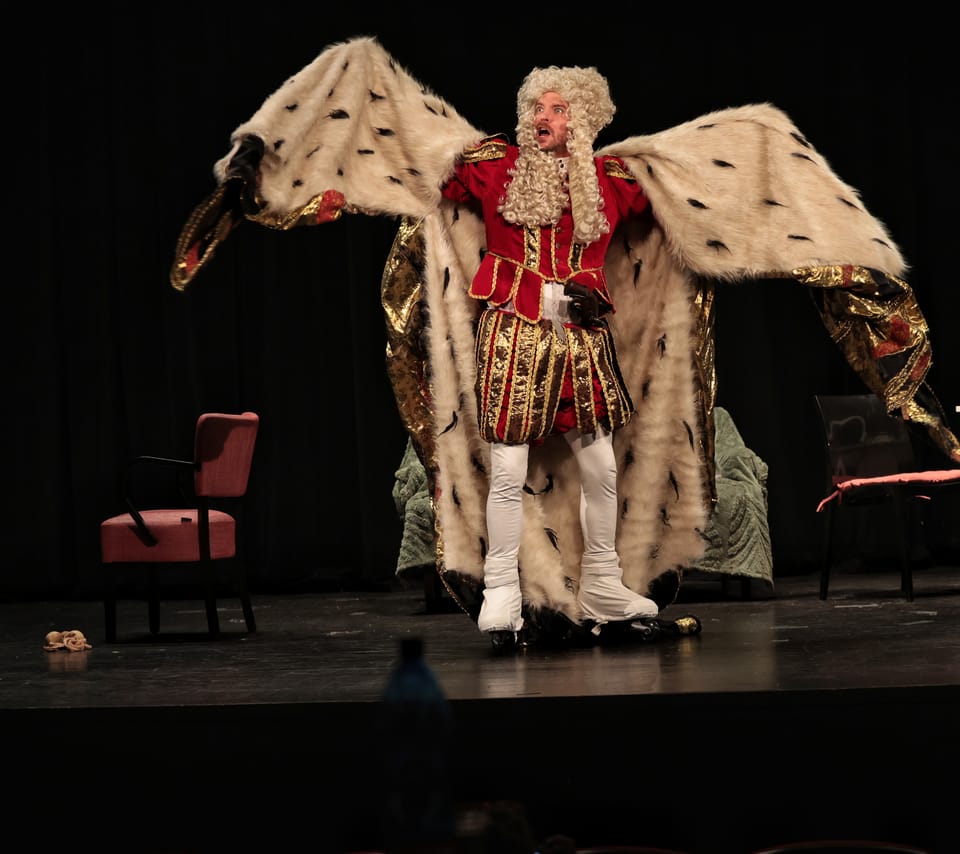 An actor in a royal robe, arms spread wide on stage