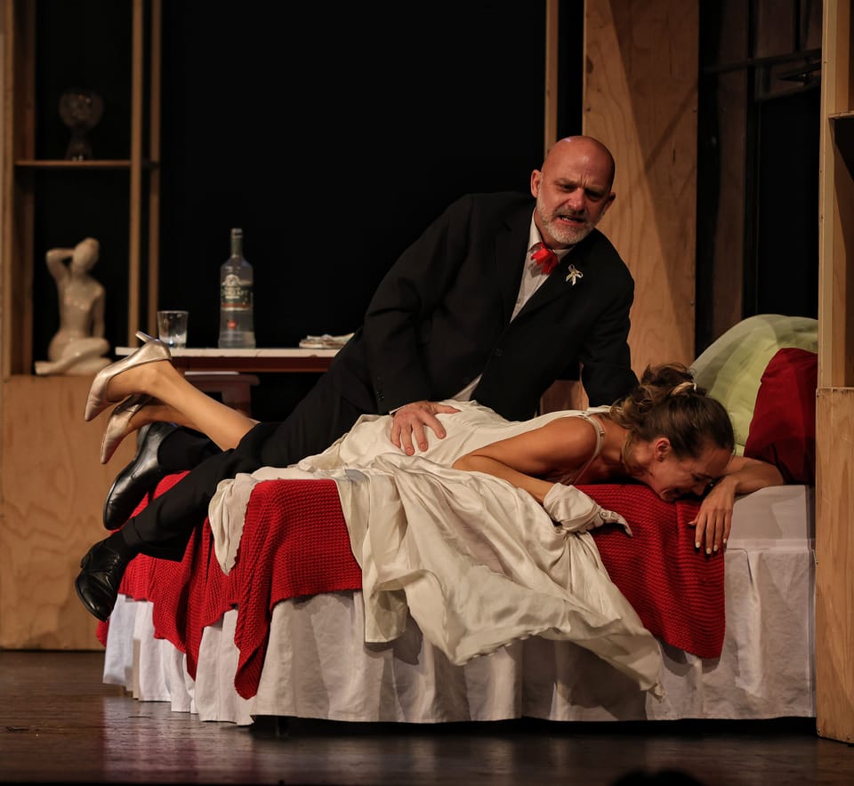 A bedroom stage scene, a man in tails leaning over a reclining actress