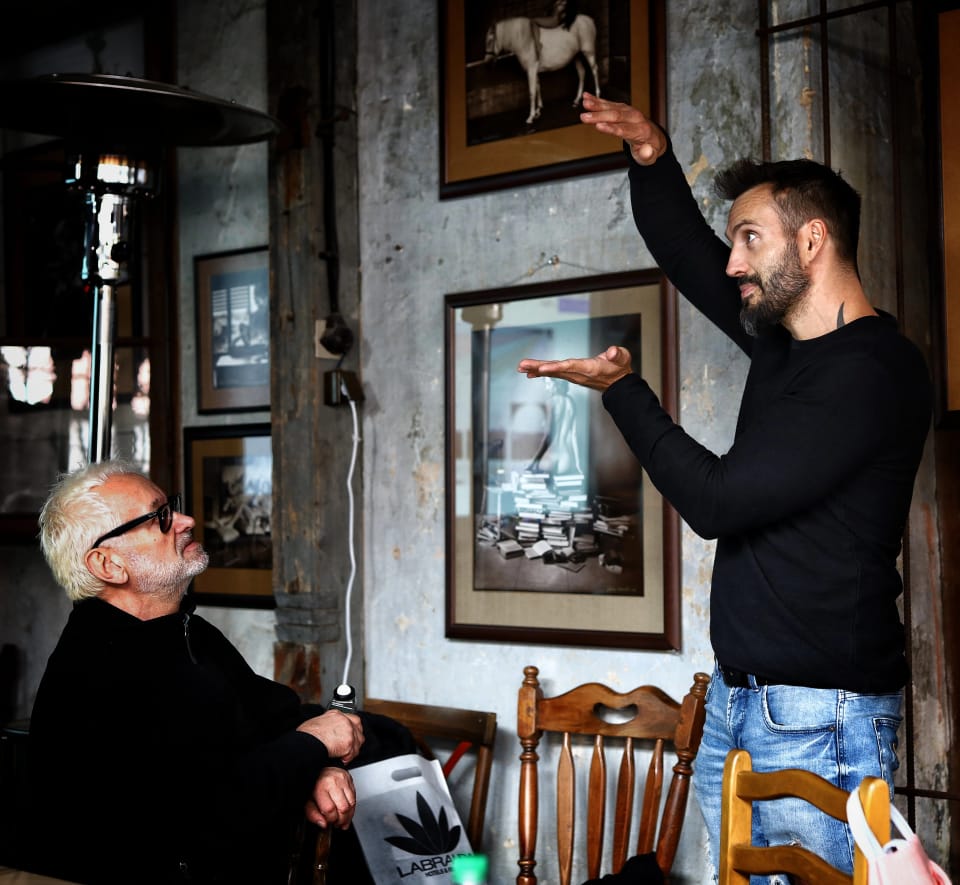 Two men in a room hung with framed photographs, one pointing at the wall