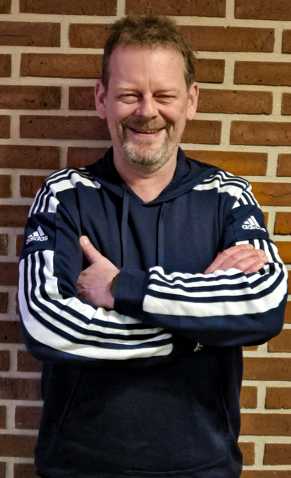 A portrait of a man with folded arms at a brick wall, a smile at the corner of his mouth