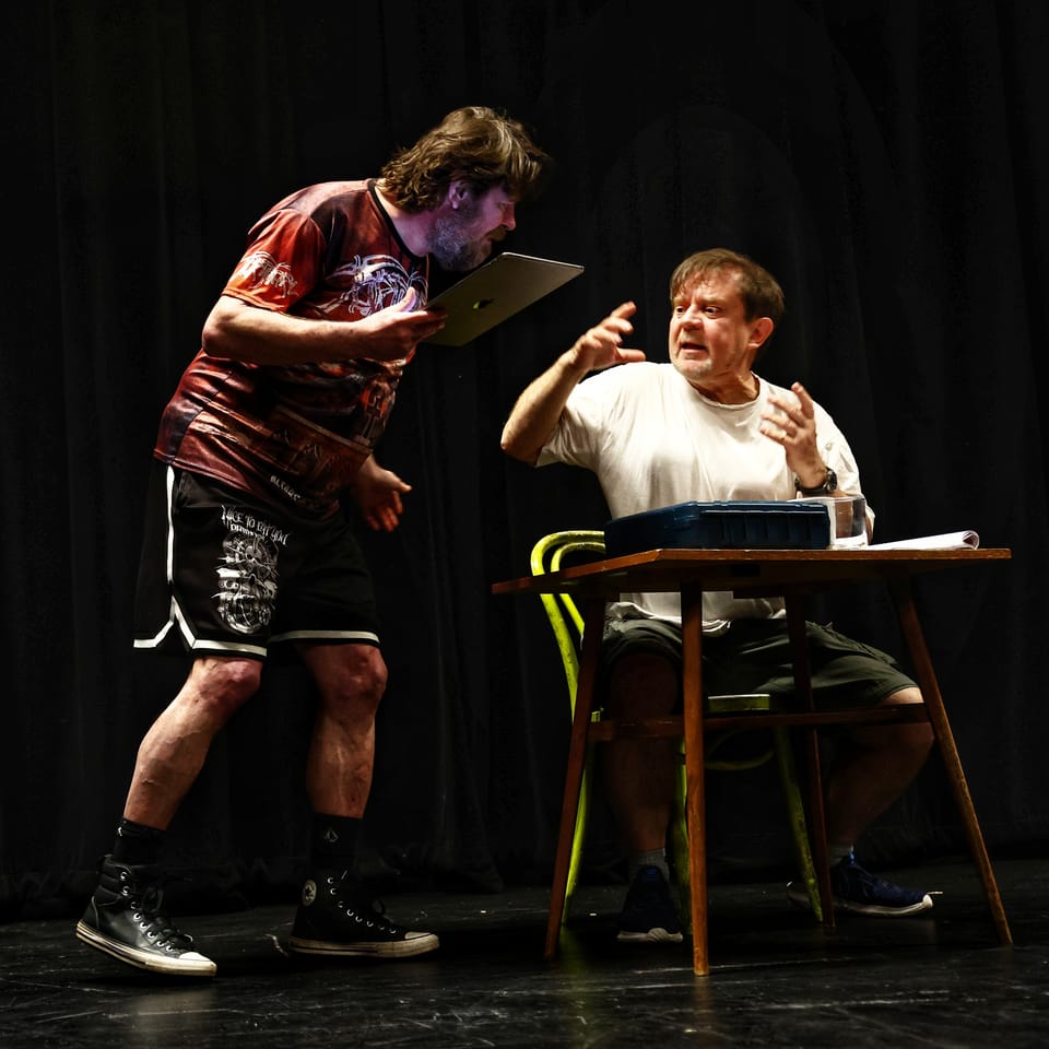 Two actors at a table with a projector, one bent over the papers