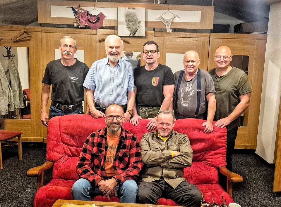 Sedm mužů v šatně, pět stojí vzadu, dva sedí na červené pohovce