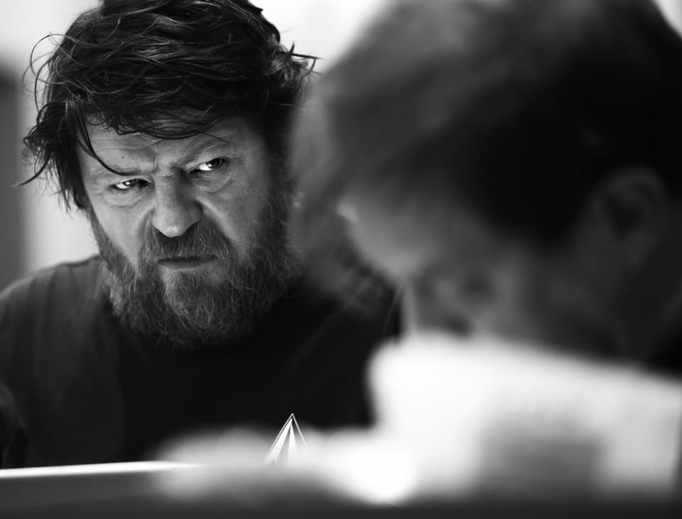 A bearded man staring fixedly across a table, the foreground blurred into white