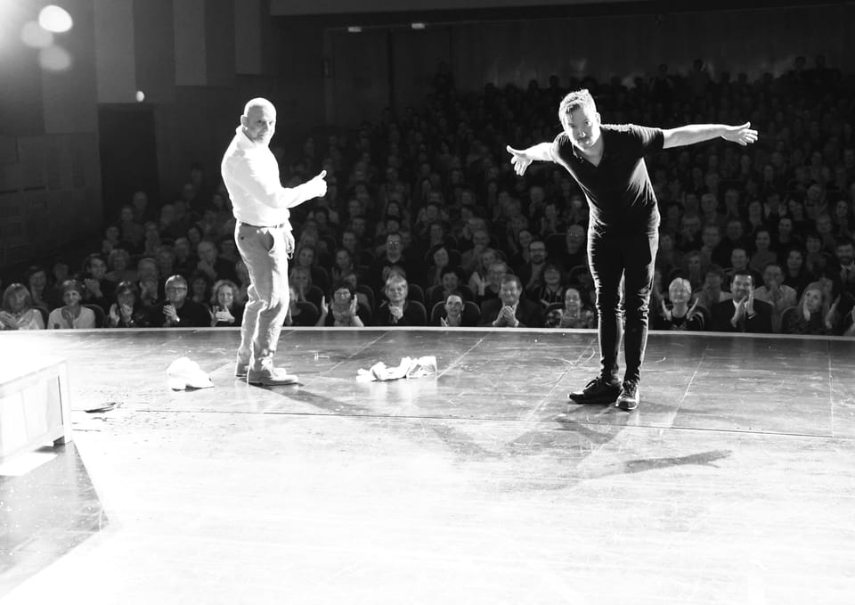 Two actors on stage facing a full house, one with arms spread