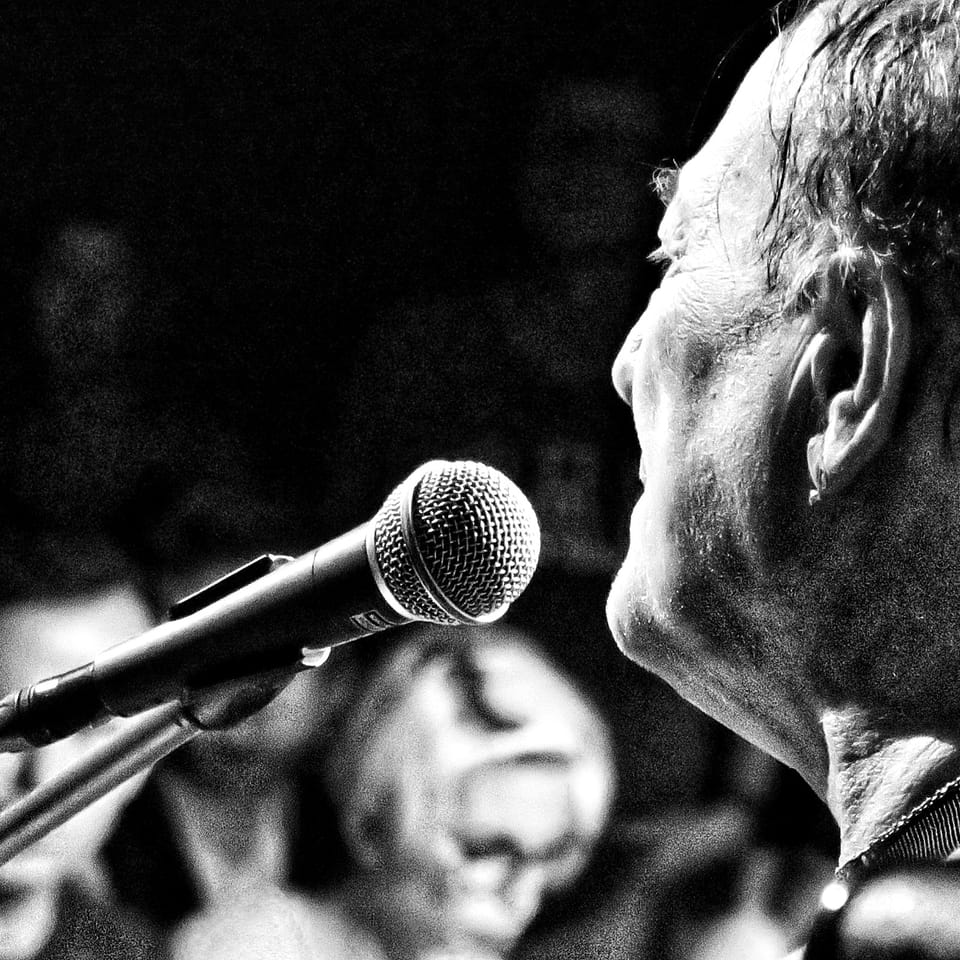 An older man in profile close to the microphone, the background dissolved into black