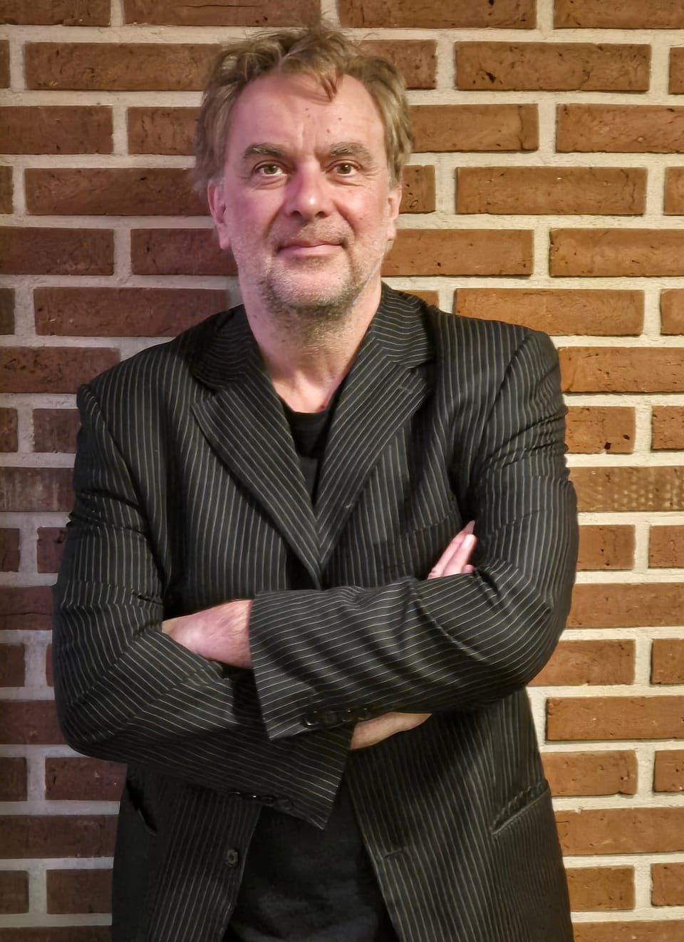 A man in a pinstriped jacket at a brick wall, looking straight into the lens