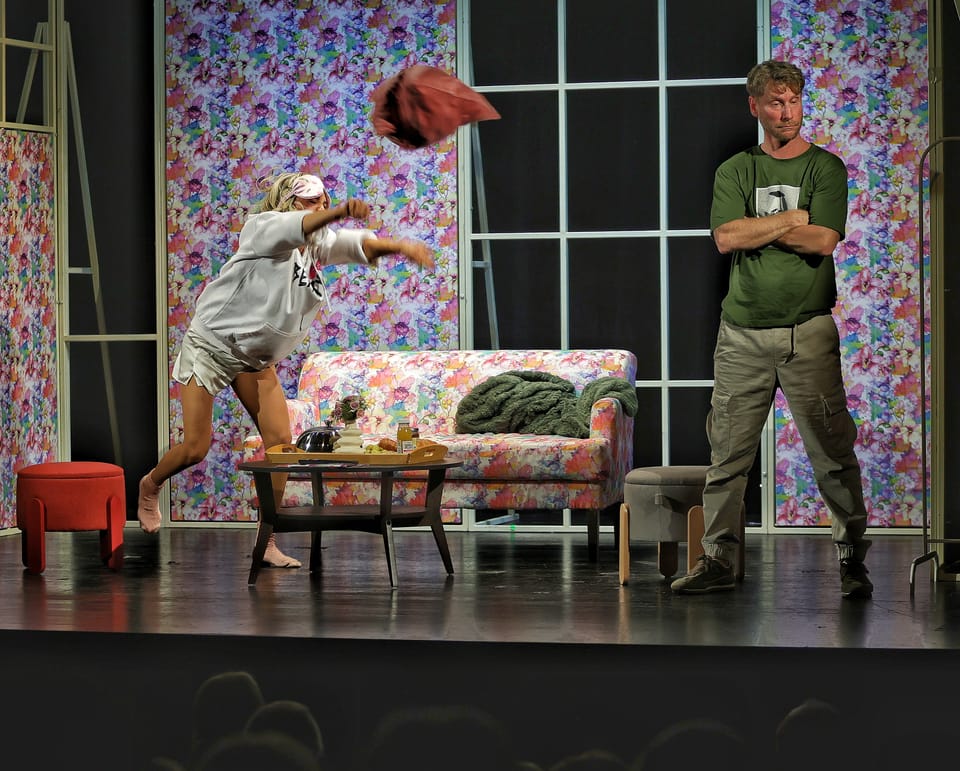 An actress vaulting the sofa in a floral-papered room while a man stands unmoved