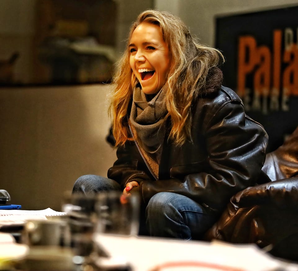 A woman in a leather jacket laughing at the table, the foreground blurred into glasses